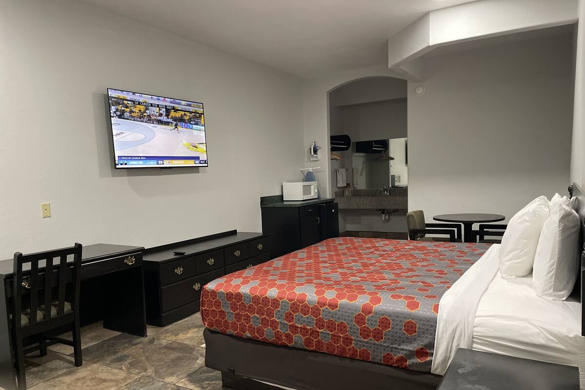 A hotel room with a bed, a desk, a chair, a TV, a small table, and a bathroom visible through an archway.