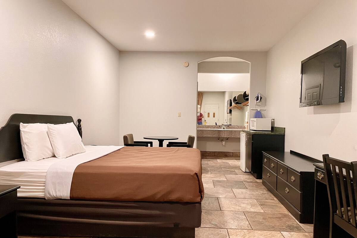A hotel room with a bed, a desk, a chair, a small table, a mirror, a TV, and a kitchenette.