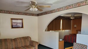 Une chambre d’hôtel avec un canapé, une chaise en bois, un lit recouvert d’une couvre-lit à motifs, une table de chevet, une lampe et un ventilateur de plafond.