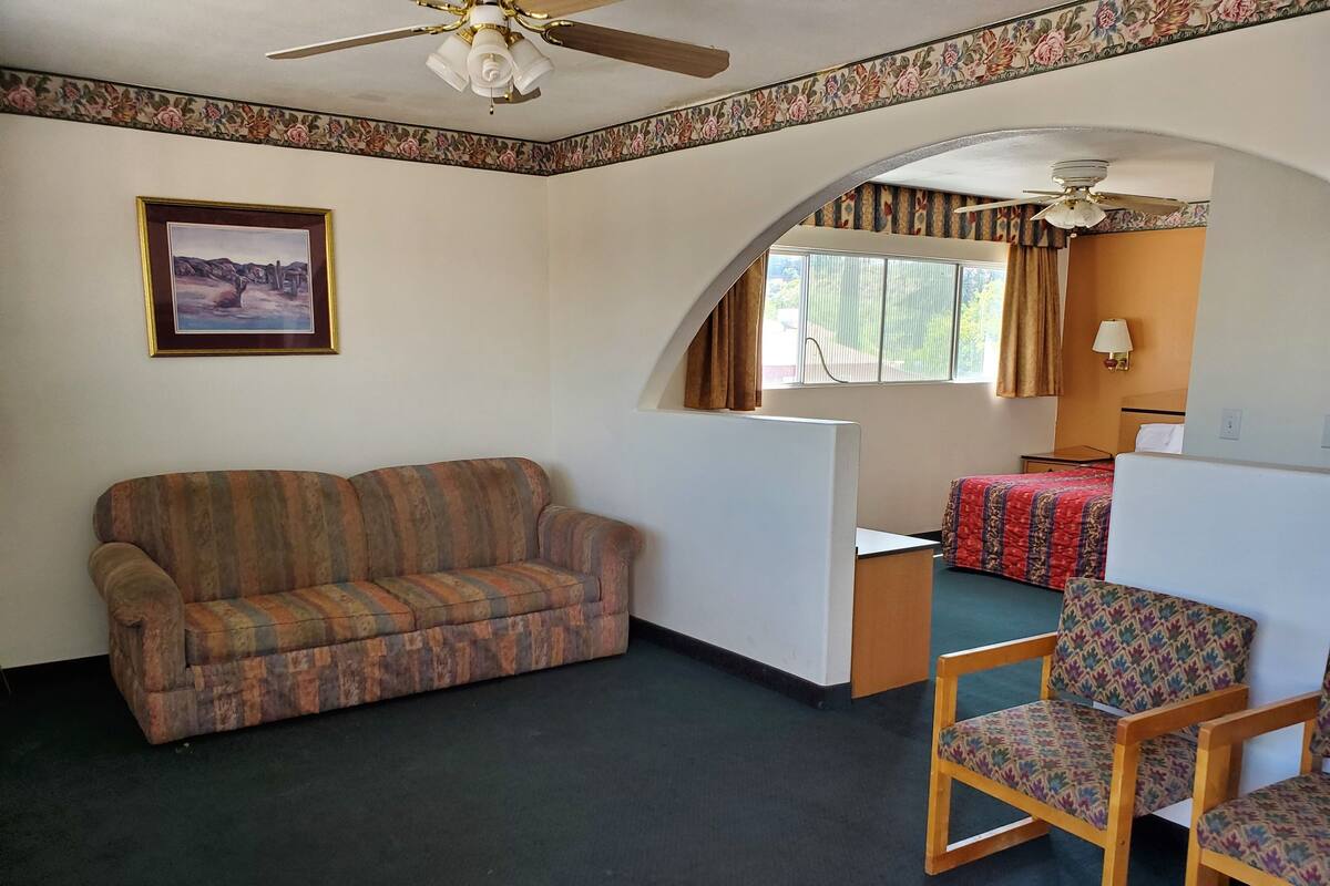 A room with a sofa, two chairs, a bed with a patterned bedspread, a ceiling fan, and a framed picture on the wall.