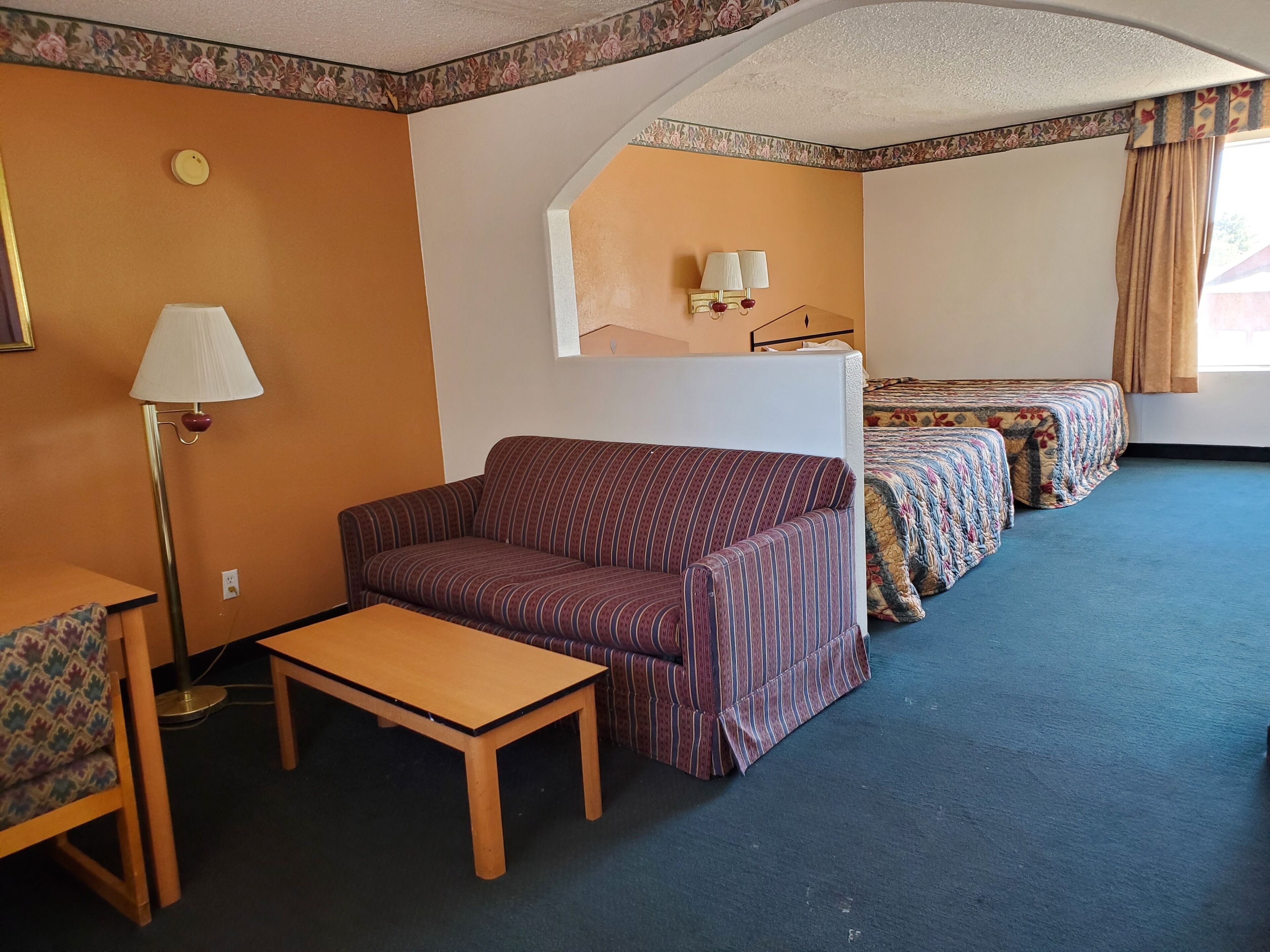 A room with a sofa, a small table, a lamp, a bed with floral bedding, and a ceiling with floral trim.