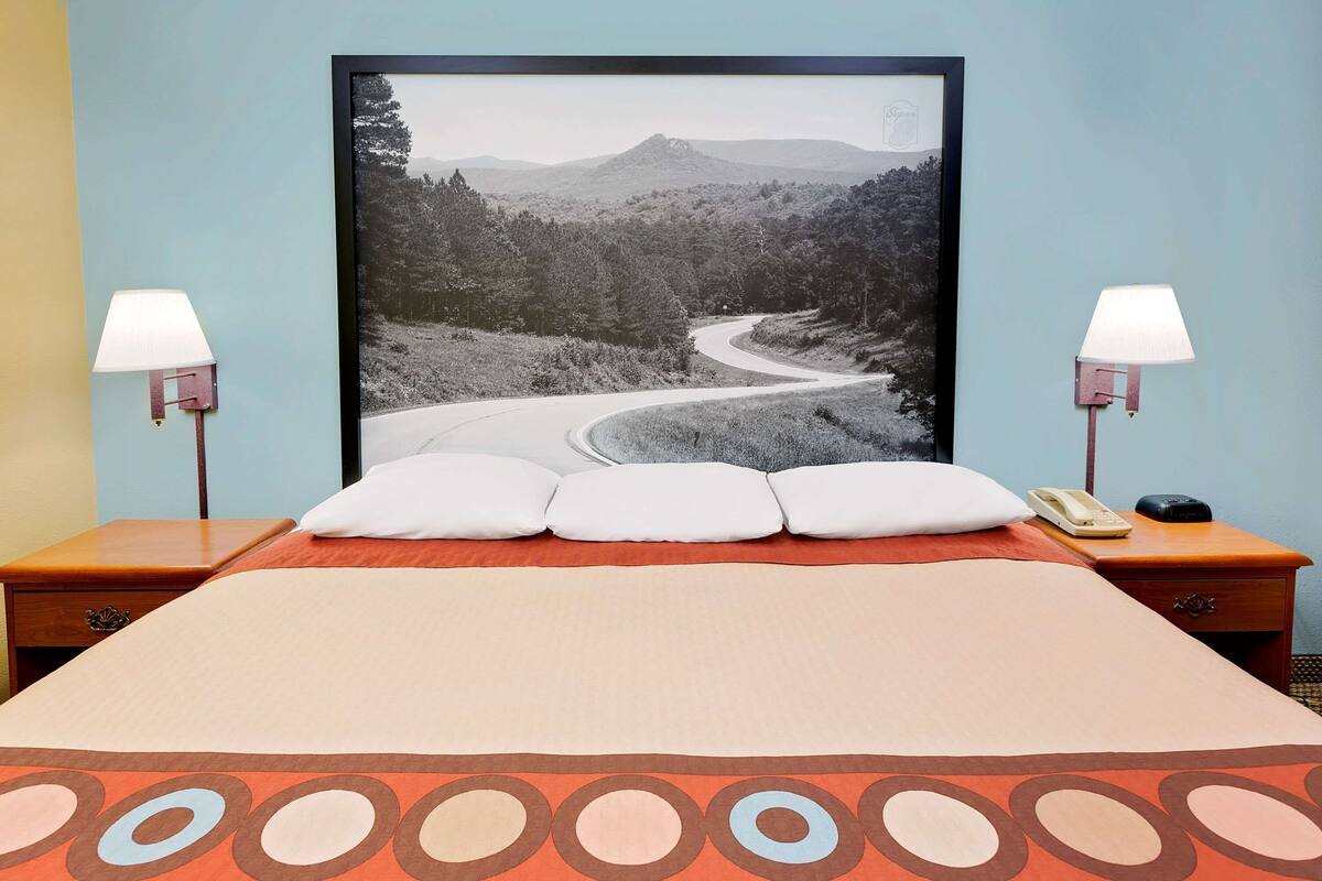 A neatly made bed with a large framed picture of a winding road and mountains, two bedside lamps, and a bedside telephone.