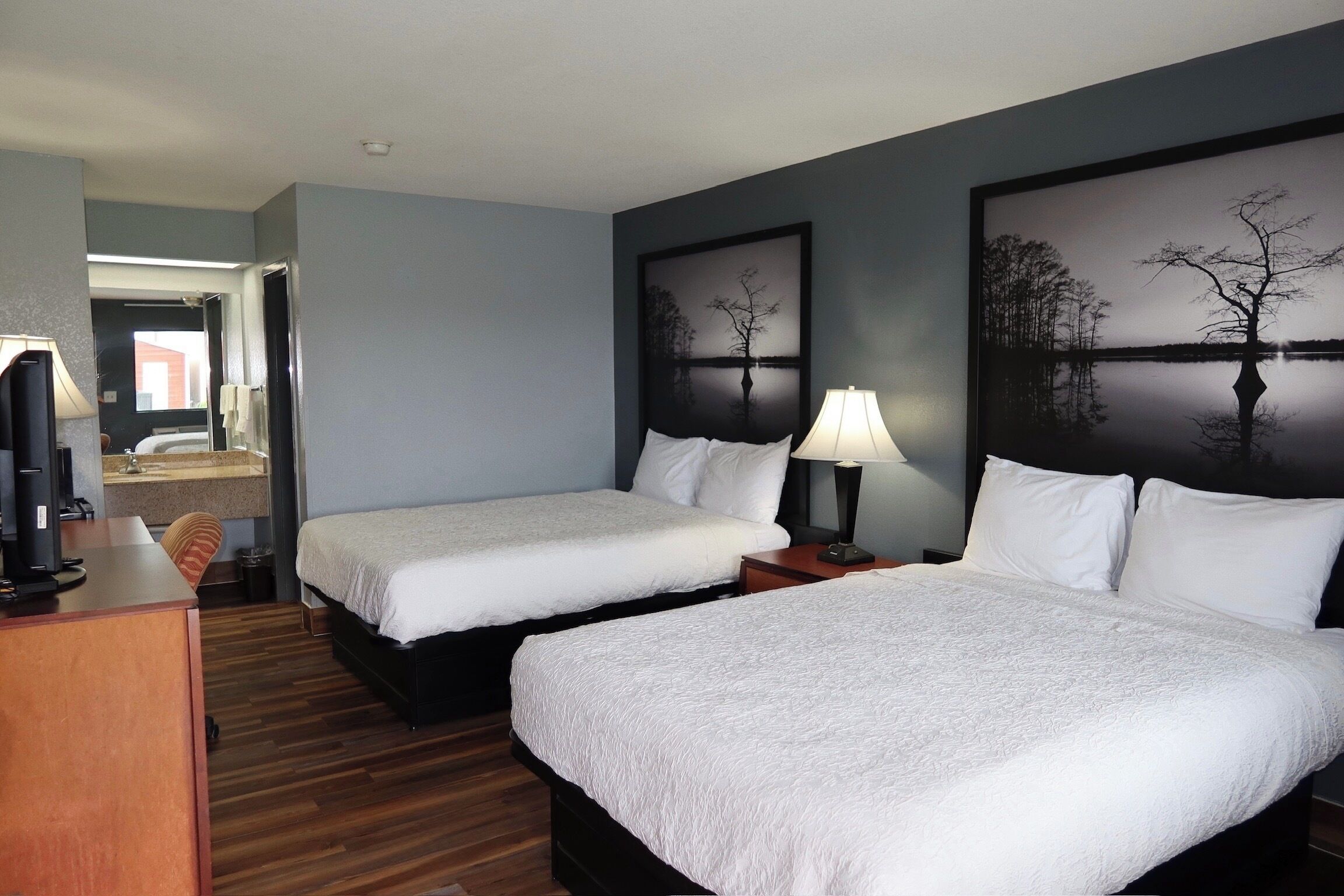 A hotel room with two beds, a desk with a computer, a lamp, and artwork on the walls.
