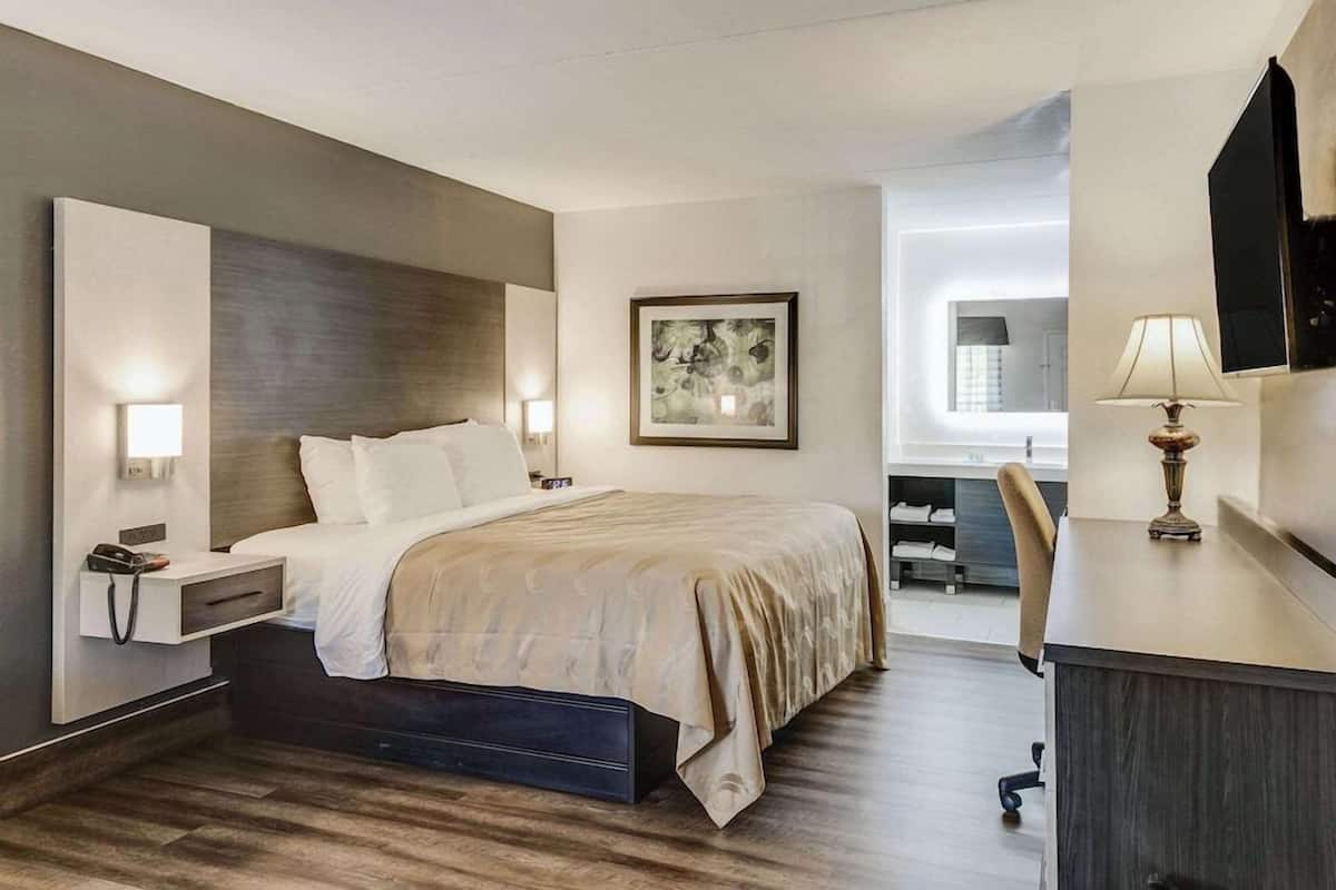 A hotel room with a bed, bedside table, lamp, desk, chair, and artwork on the wall.