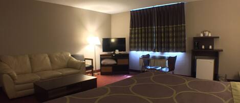 A hotel room with a sofa, a small table, a TV, a desk with a chair, and a refrigerator.