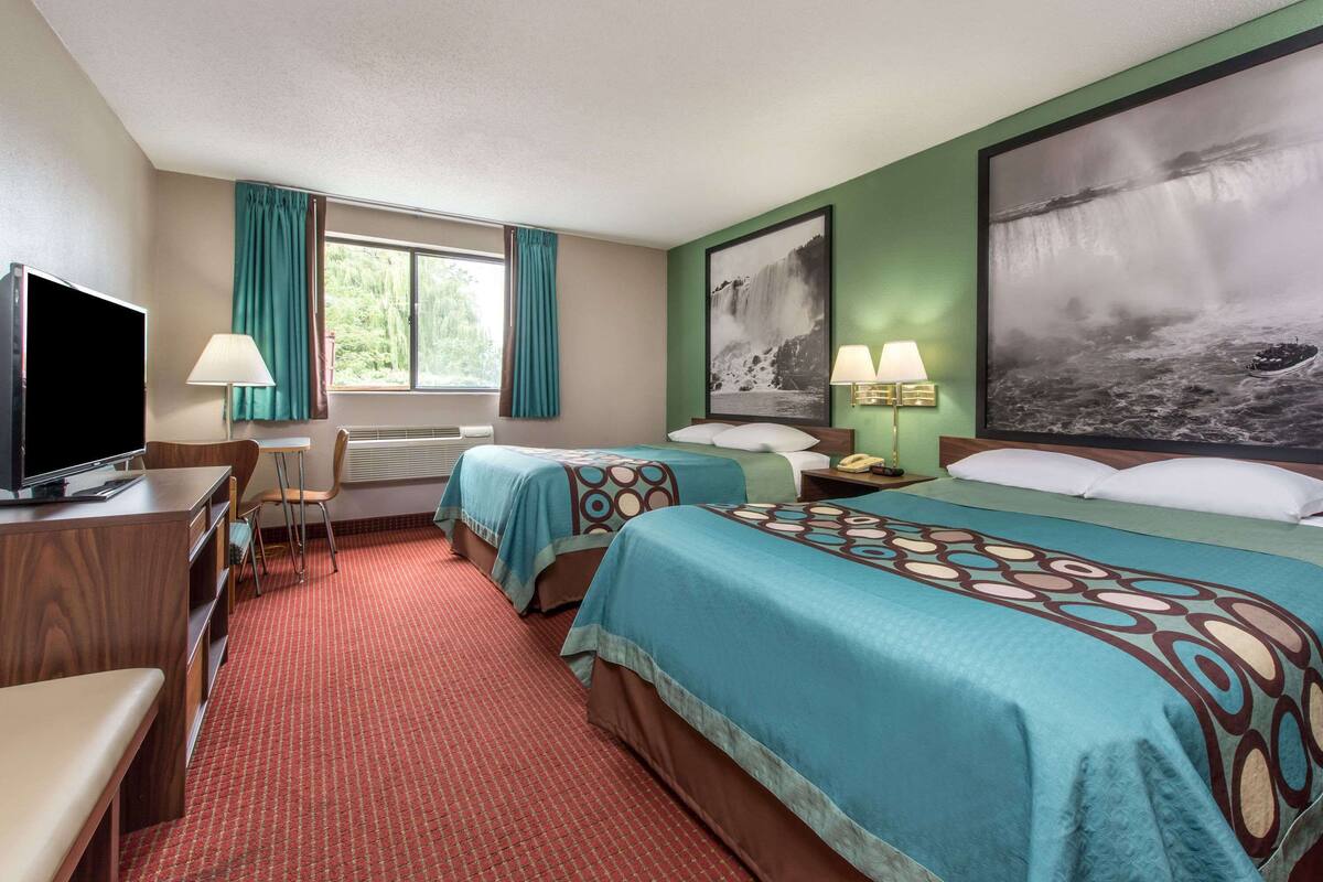 Ein Hotelzimmer mit zwei Betten, einem Schreibtisch mit Fernseher, einem Sessel, einem Fenster mit Vorhängen und zwei Wandlampen.