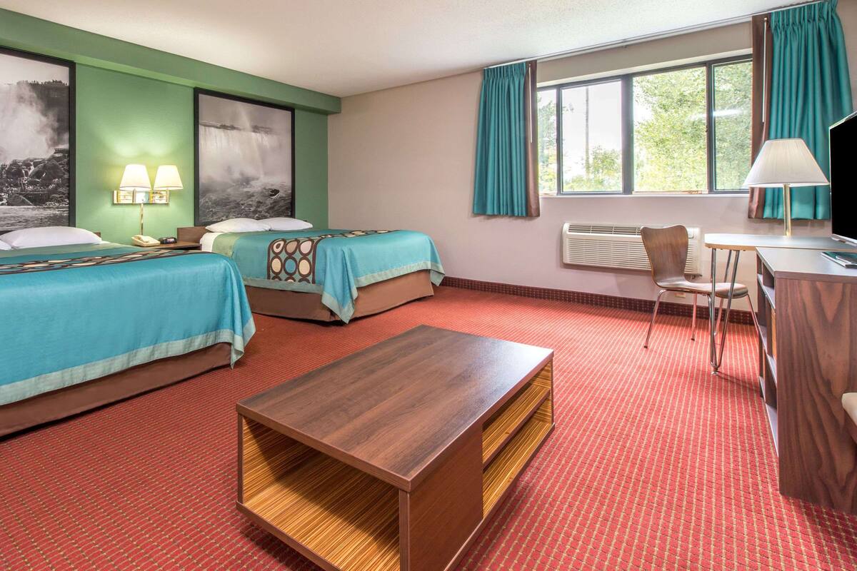 Ein Hotelzimmer mit zwei Betten, einem Holztisch, einem Schreibtisch mit Stuhl und einem Fenster mit Vorhängen.
