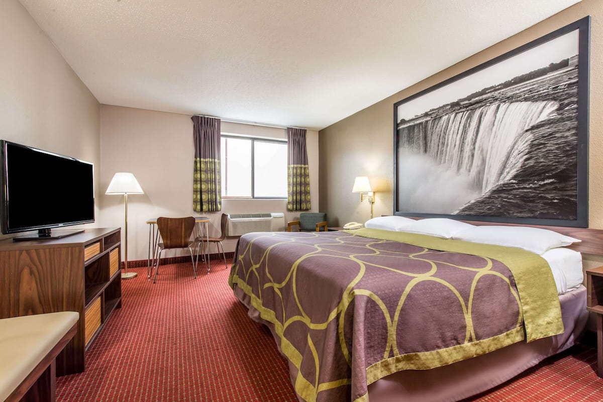 Ein Hotelzimmer mit einem großen Bett, einem Flachbildfernseher, einem Schreibtisch mit Stuhl, einem Fenster mit Vorhängen und einem Wandbild mit einem Wasserfall.