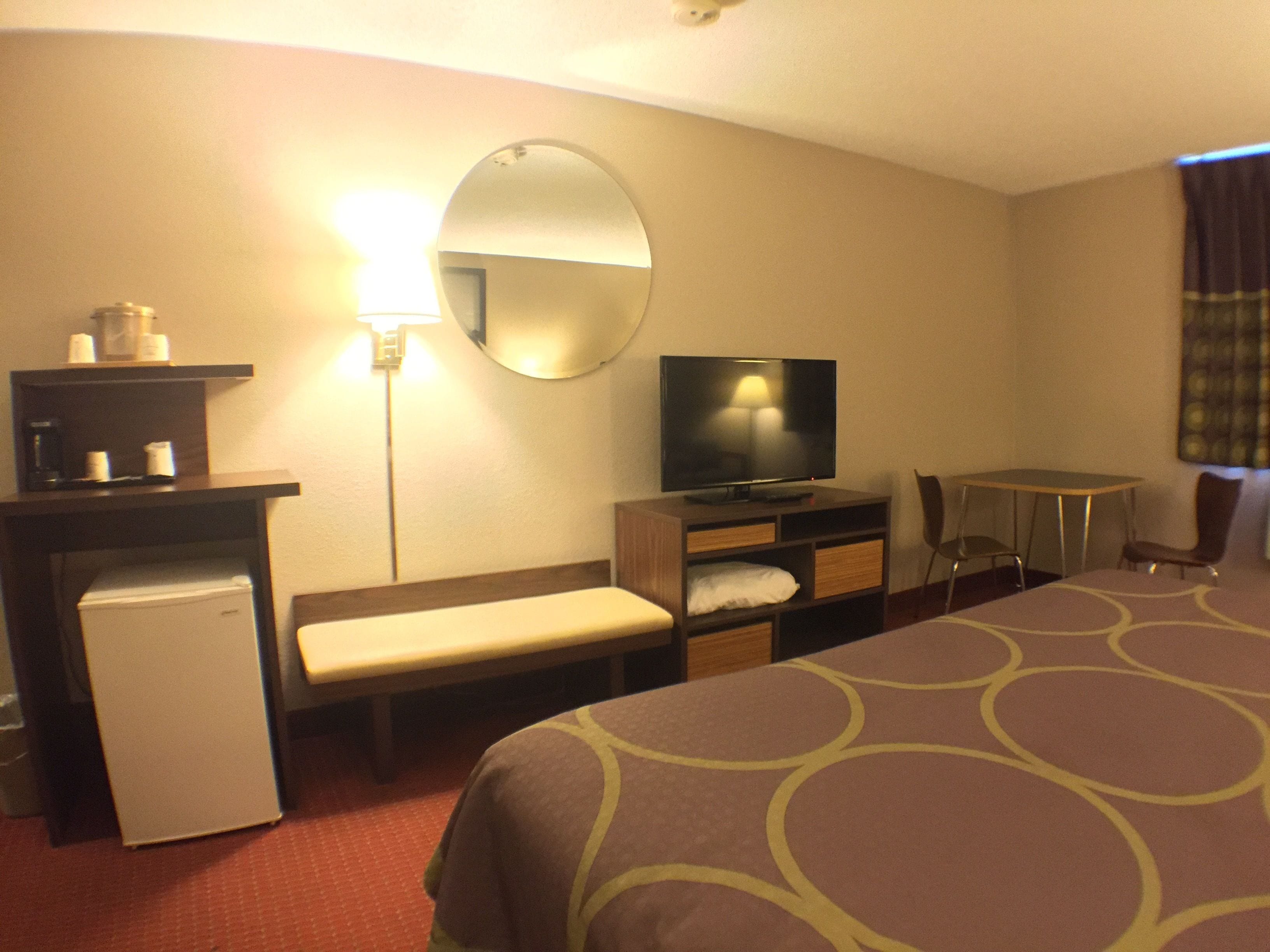 Una camera d'albergo con un letto, una scrivania, una televisione, un piccolo frigorifero, uno specchio e una lampada.