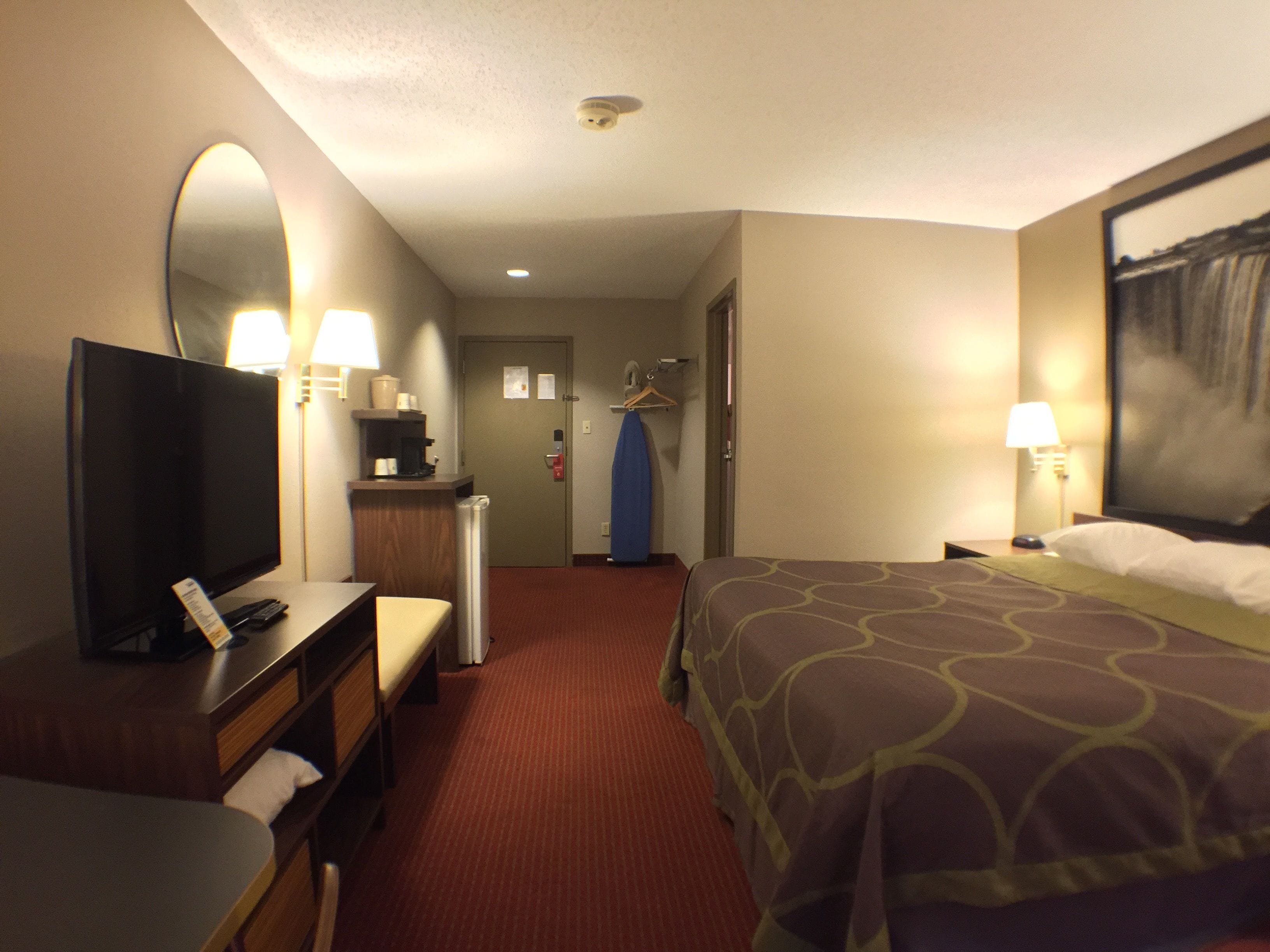 A hotel room with a bed, a desk with a computer, a mirror, a coat rack, and a door leading to a hallway.