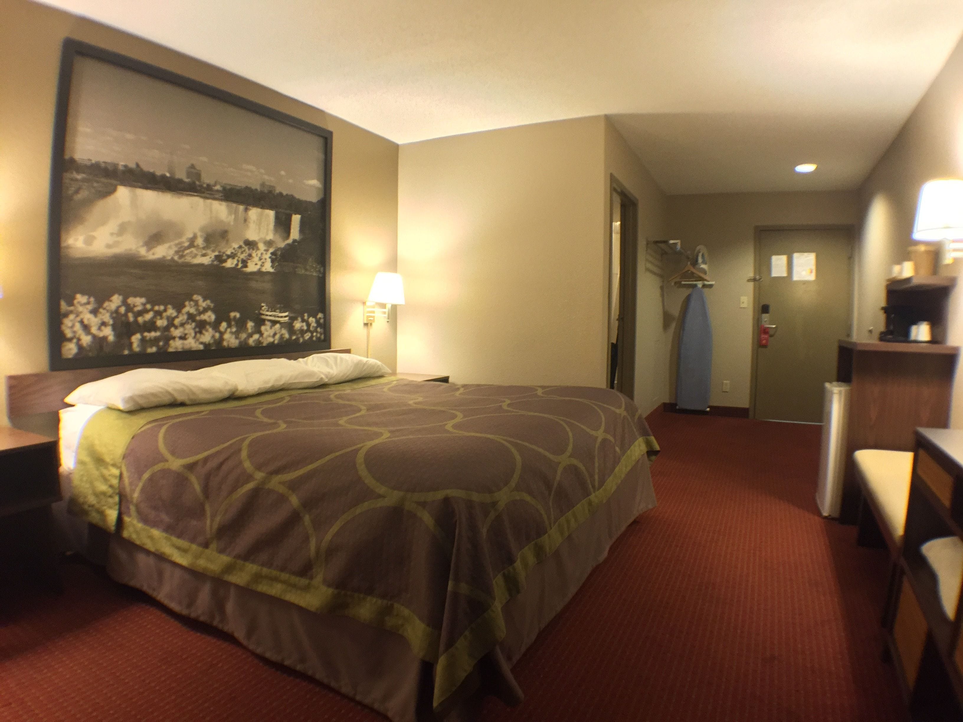 A hotel room with a large bed, a nightstand, a lamp, a painting on the wall, and a door leading to a hallway.