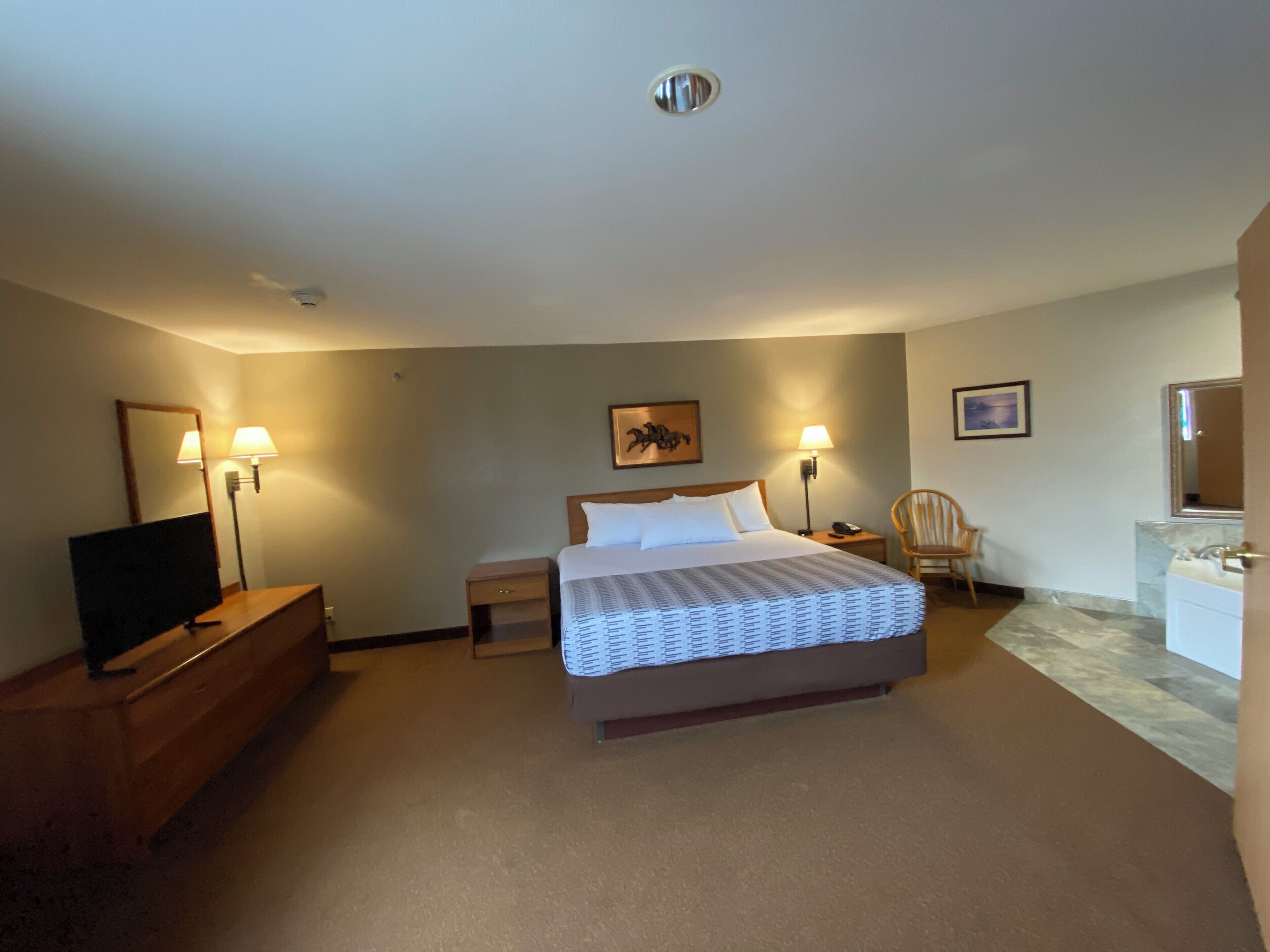 A hotel room with a bed, bedside lamps, a chair, a desk with a TV, and a bathroom with a mirror.