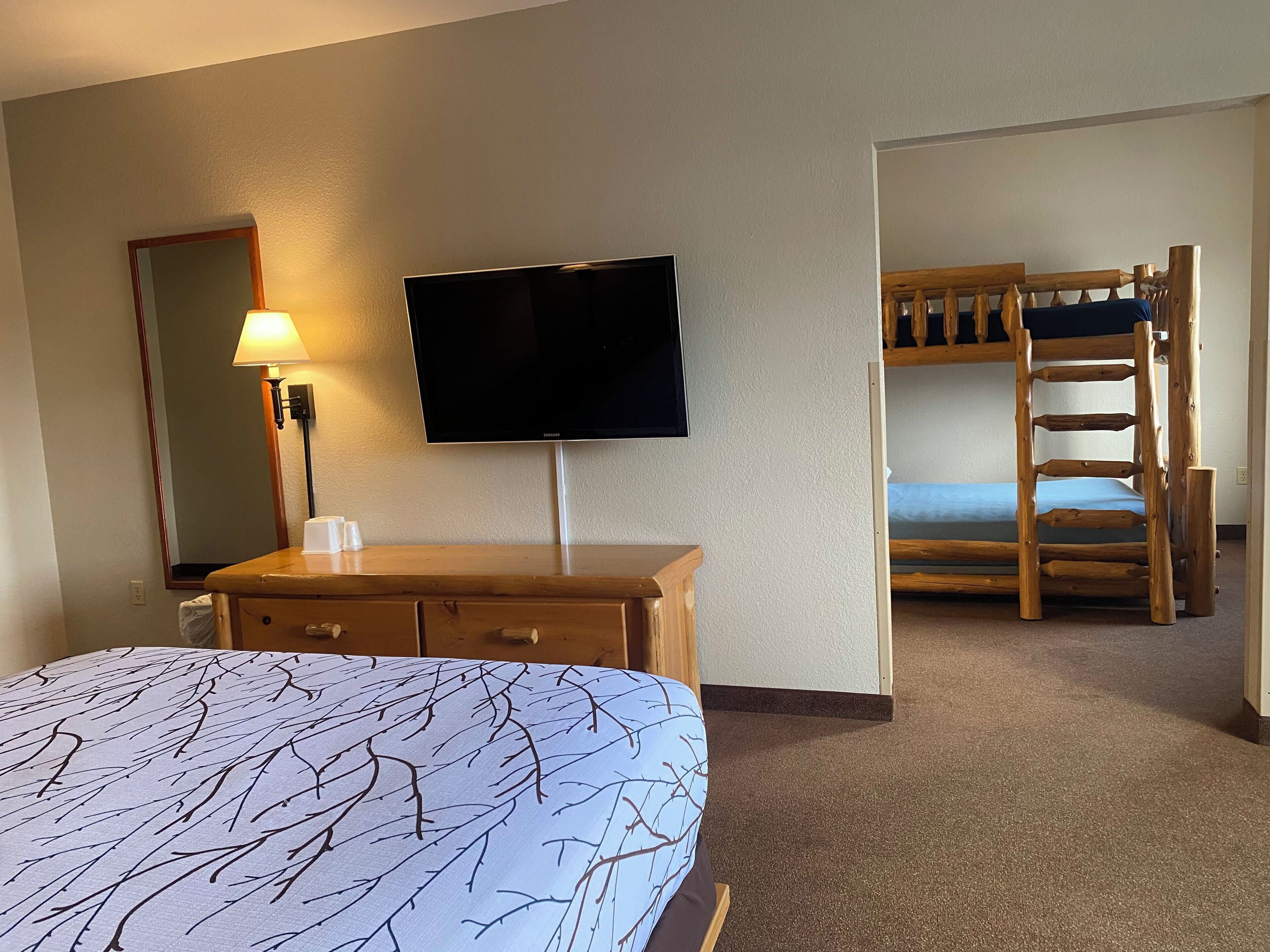 A hotel room with a bed, a TV, a wooden dresser, a mirror, and a bunk bed.