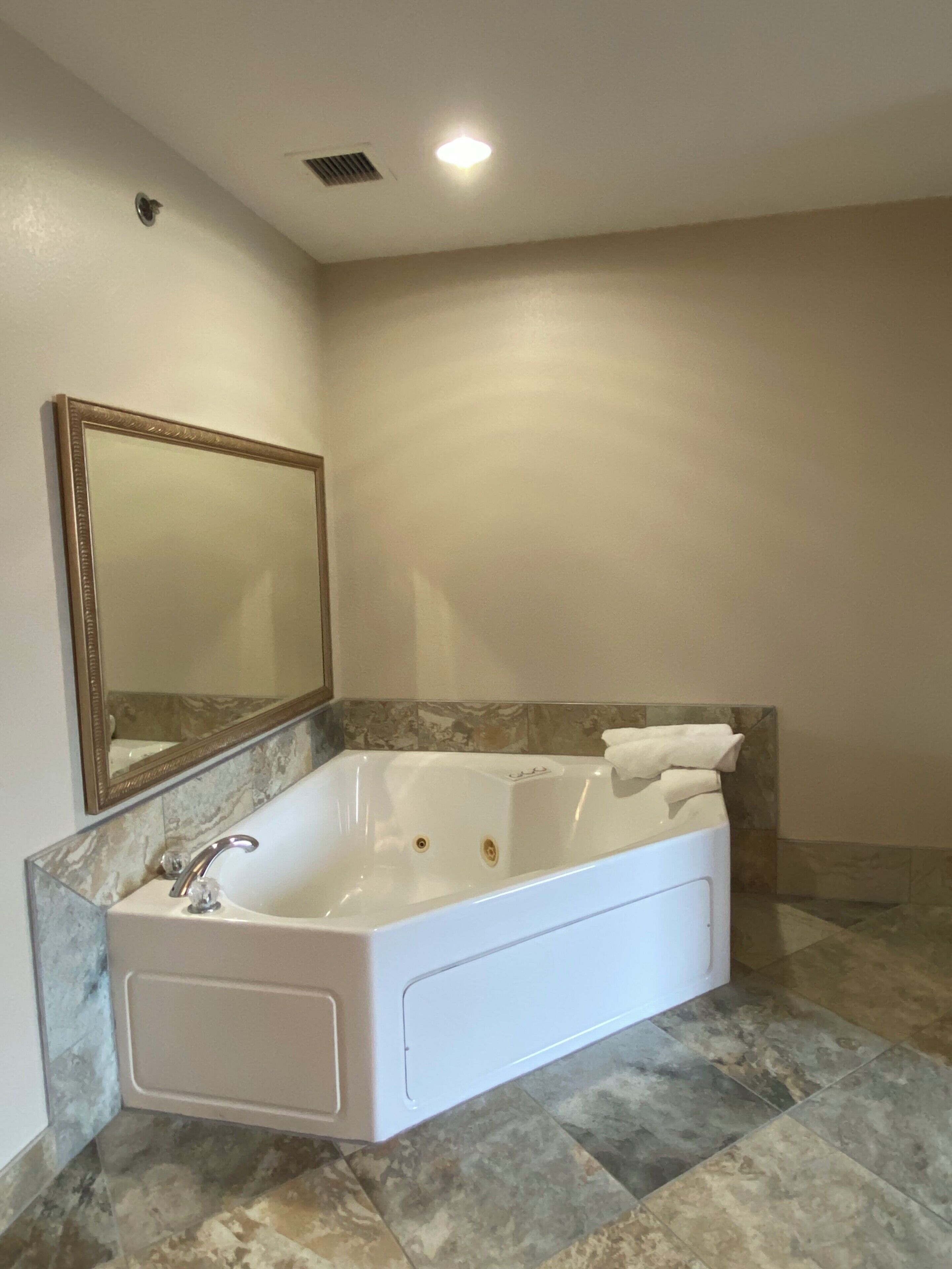 A bathroom with a large bathtub, a wall-mounted mirror, and a marble-tiled floor.