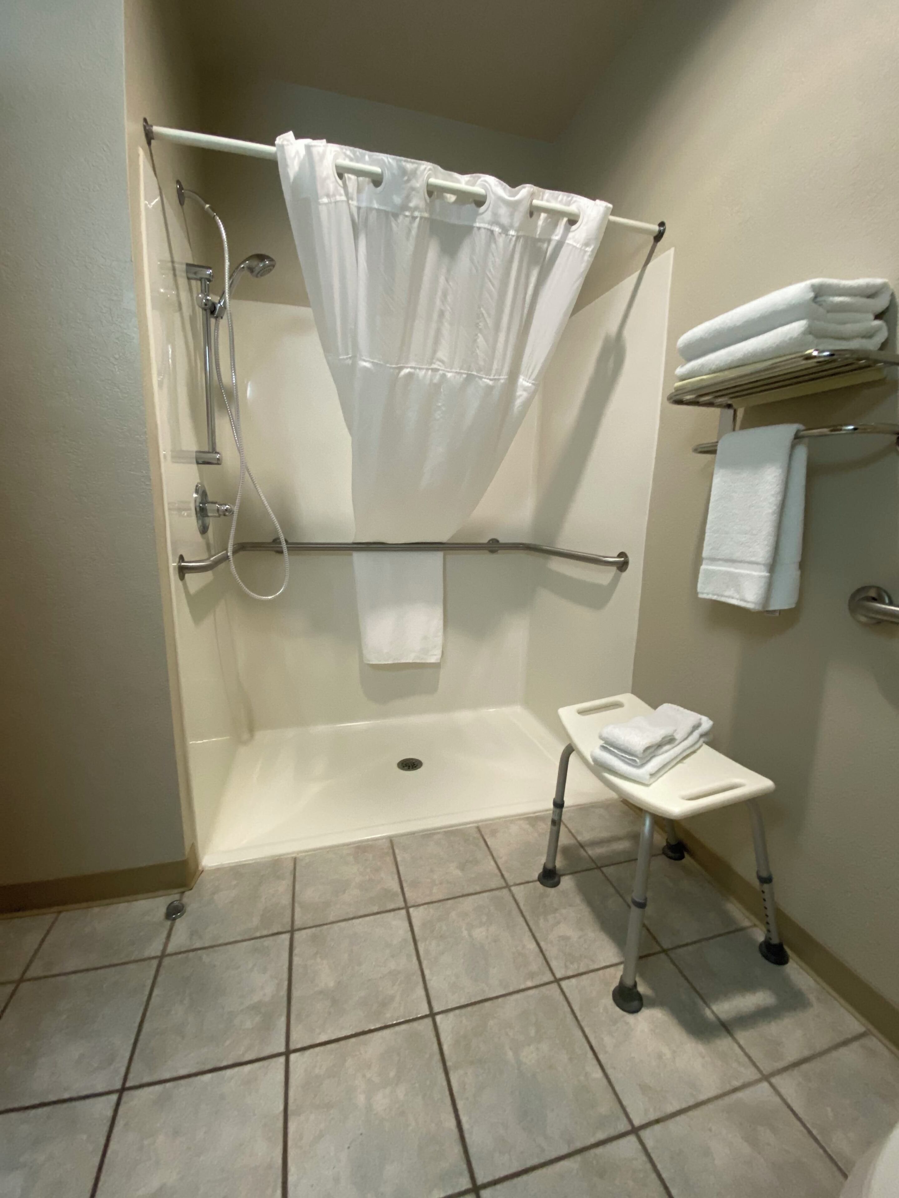 A shower with a white curtain, a small step stool, and a towel rack with folded towels.