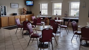Un espace repas comprenant plusieurs tables et chaises, un téléviseur fixé au mur et un coin cuisine avec des meubles en bois.