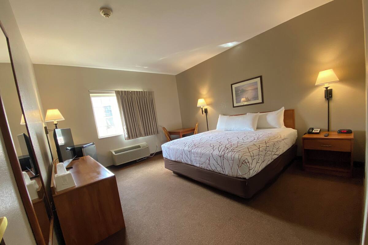 A hotel room with a bed, bedside lamps, a desk, a chair, a heater, a window with curtains, and a painting on the wall.