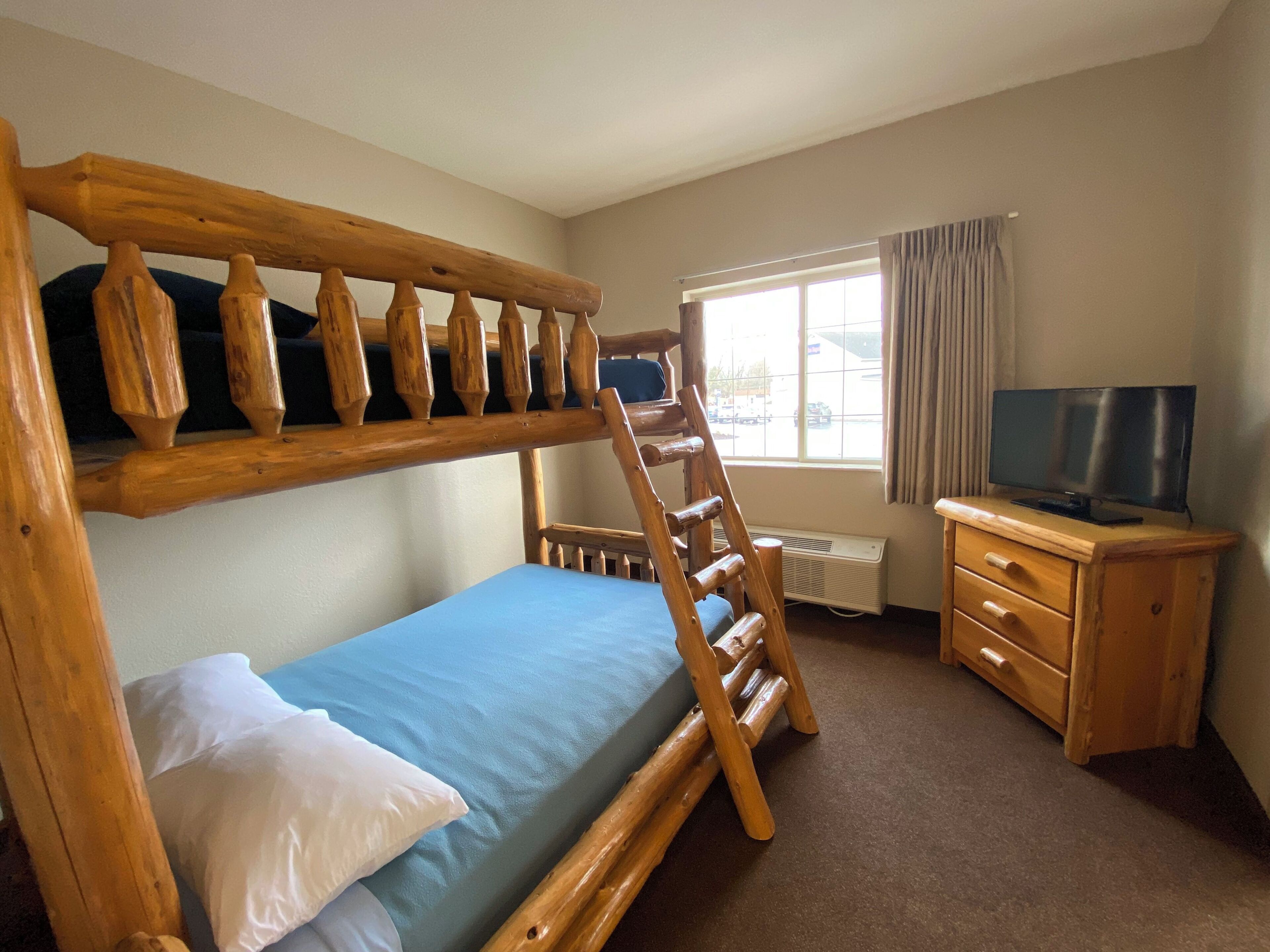 A bunk bed with a wooden ladder, a single bed with a blue mattress, a wooden dresser with a television, and a window with curtains.