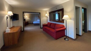 Une chambre d’hôtel avec un canapé rouge, un bureau en bois, un téléviseur à écran plat, un grand miroir et un lit à l’arrière-plan.