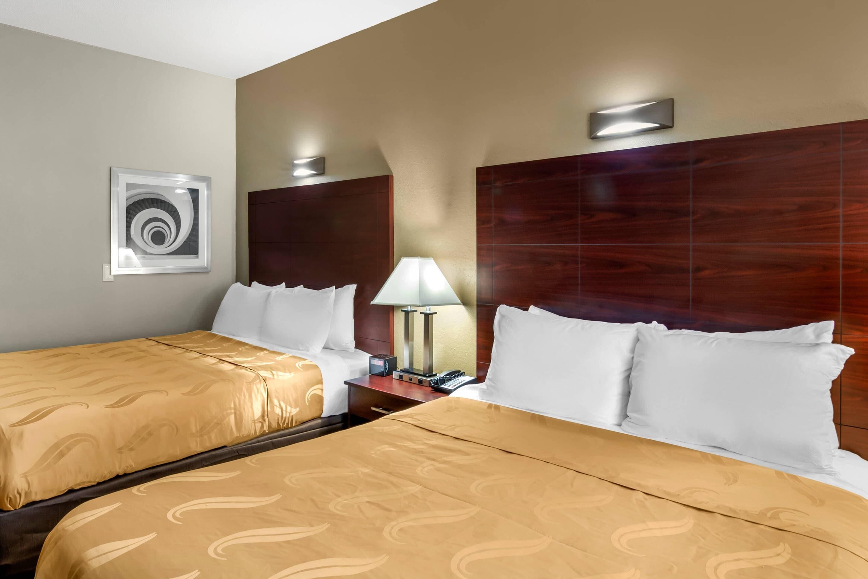 A hotel room with two beds, a nightstand, a lamp, and a wall-mounted light fixture.