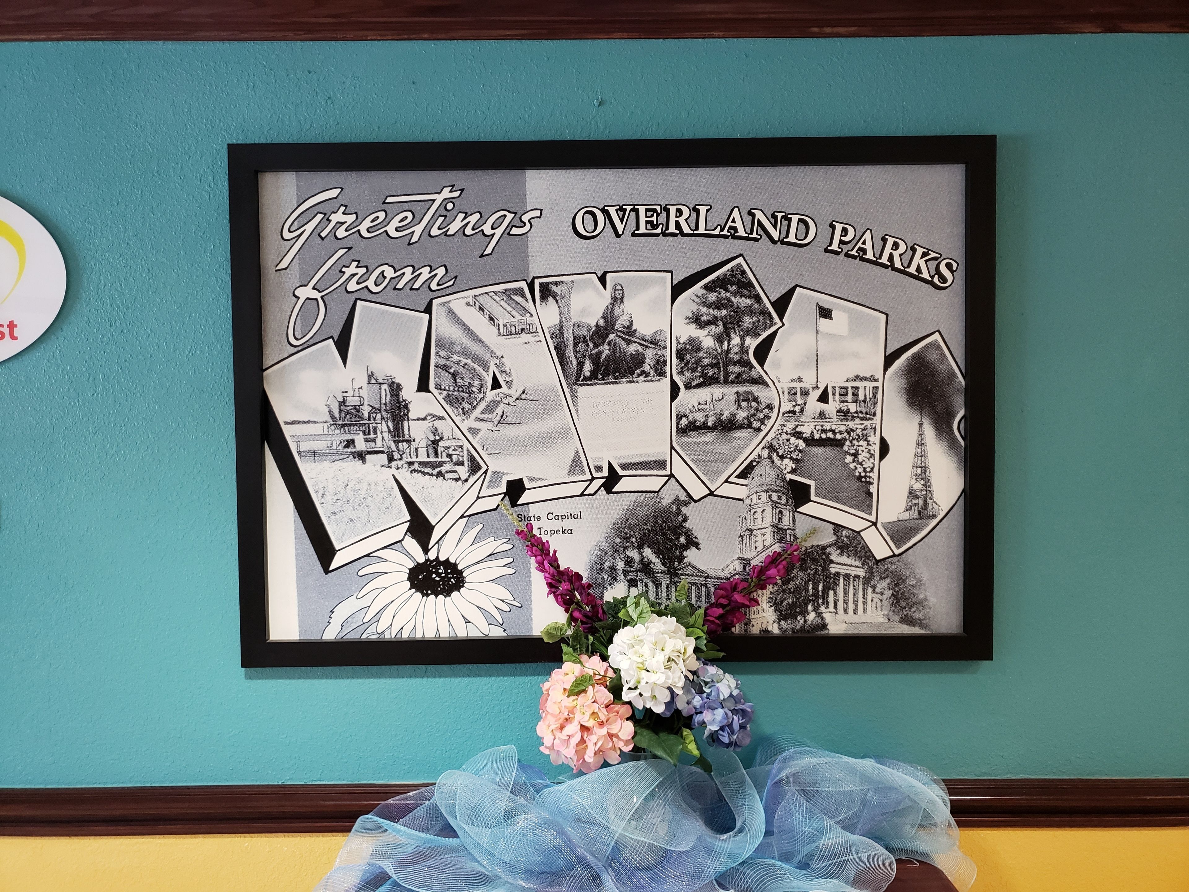 Uma colagem emoldurada intitulada 'Greetings from Overland Parks' com diversas fotografias em preto e branco de pontos turísticos e um vaso de flores.