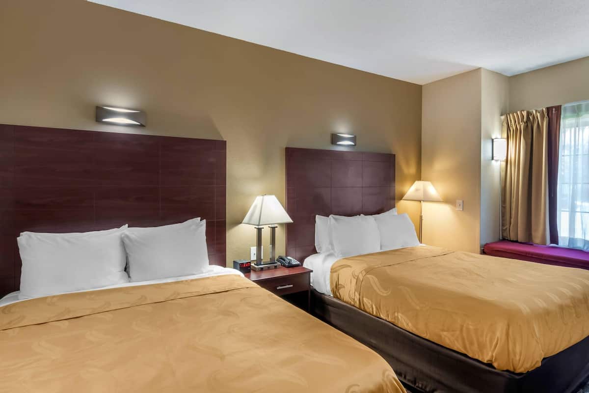 Una habitación de hotel con dos camas, una mesita de noche con lámpara, una ventana con cortinas y un aplique de pared.
