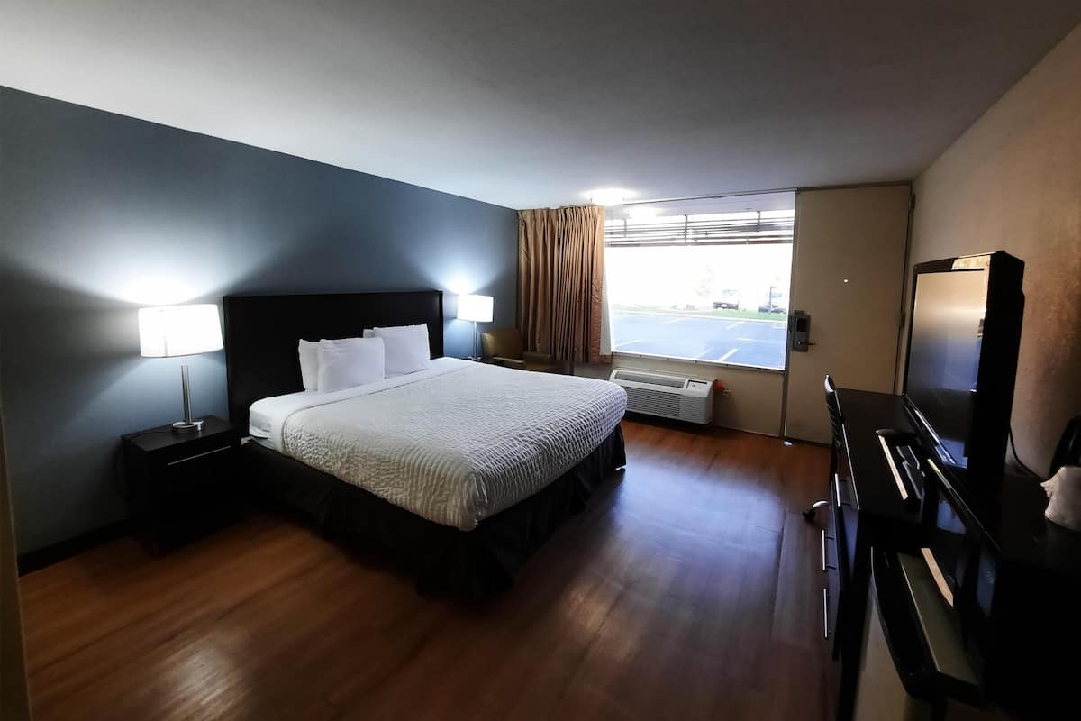 A hotel room with a bed, bedside lamps, a desk, a chair, a large window with curtains, and a flat-screen TV.