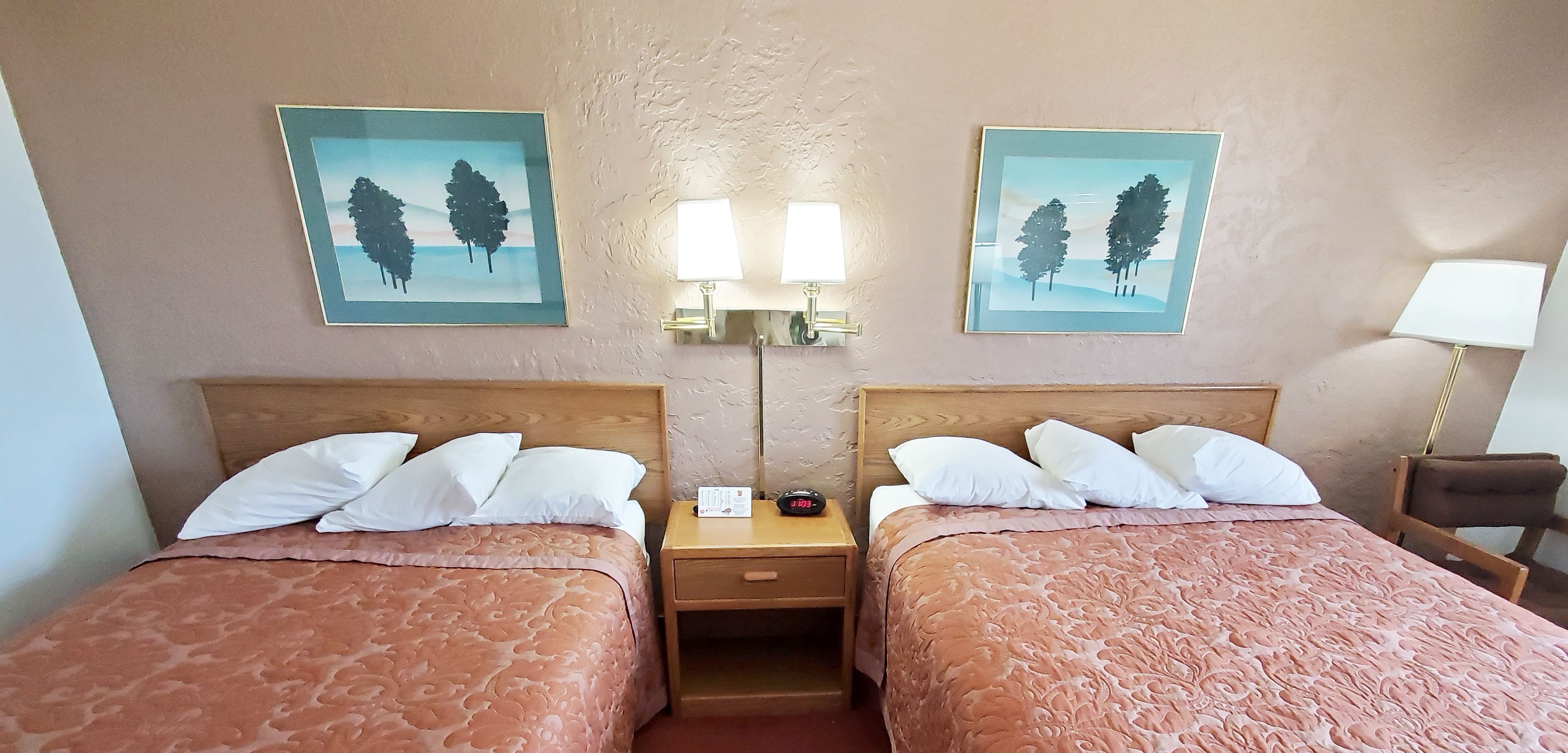 A hotel room with two beds, each with a wooden headboard and a patterned bedspread. There are two wall-mounted lamps and two framed pictures of trees.