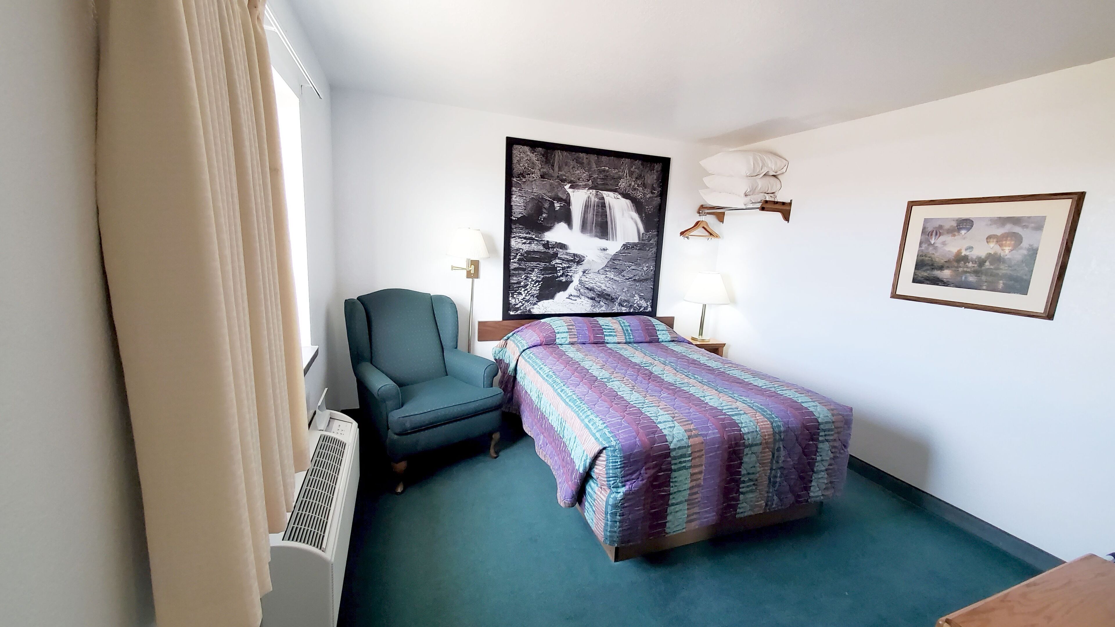 A hotel room with a bed, a chair, a radiator, a wall-mounted lamp, and a framed picture.