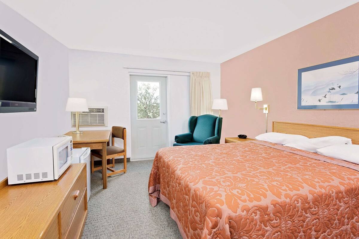 A hotel room with a bed, a desk with a microwave, a chair, a television, and a painting on the wall.