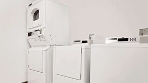 A laundry room with two front-loading washing machines and a dryer, a control panel, and a small shelf with items. - Super 8 by Wyndham Cut Bank (Cut Bank)