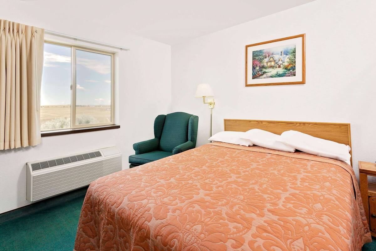 A hotel room with a large bed, a chair, a window with curtains, a radiator, a wall-mounted lamp, and a framed picture.