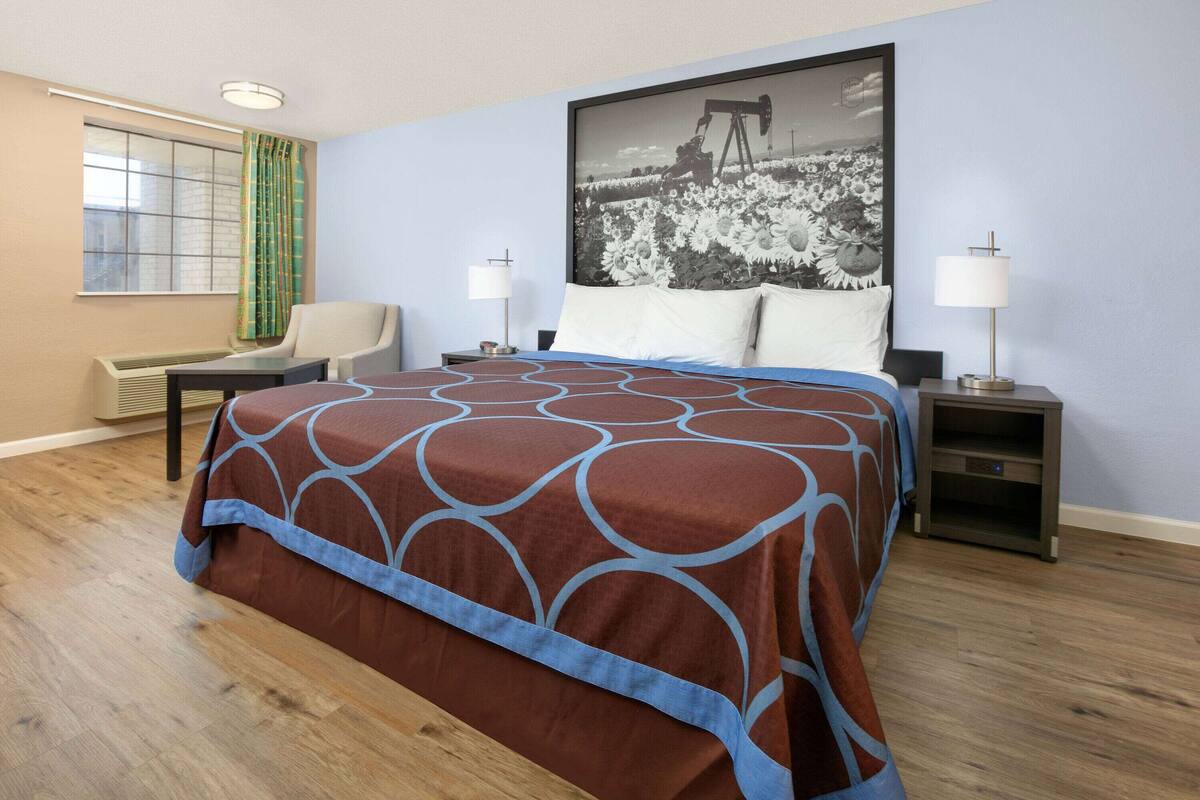 A hotel room with a large bed, two bedside tables, a chair, a window with curtains, and a wall-mounted picture.
