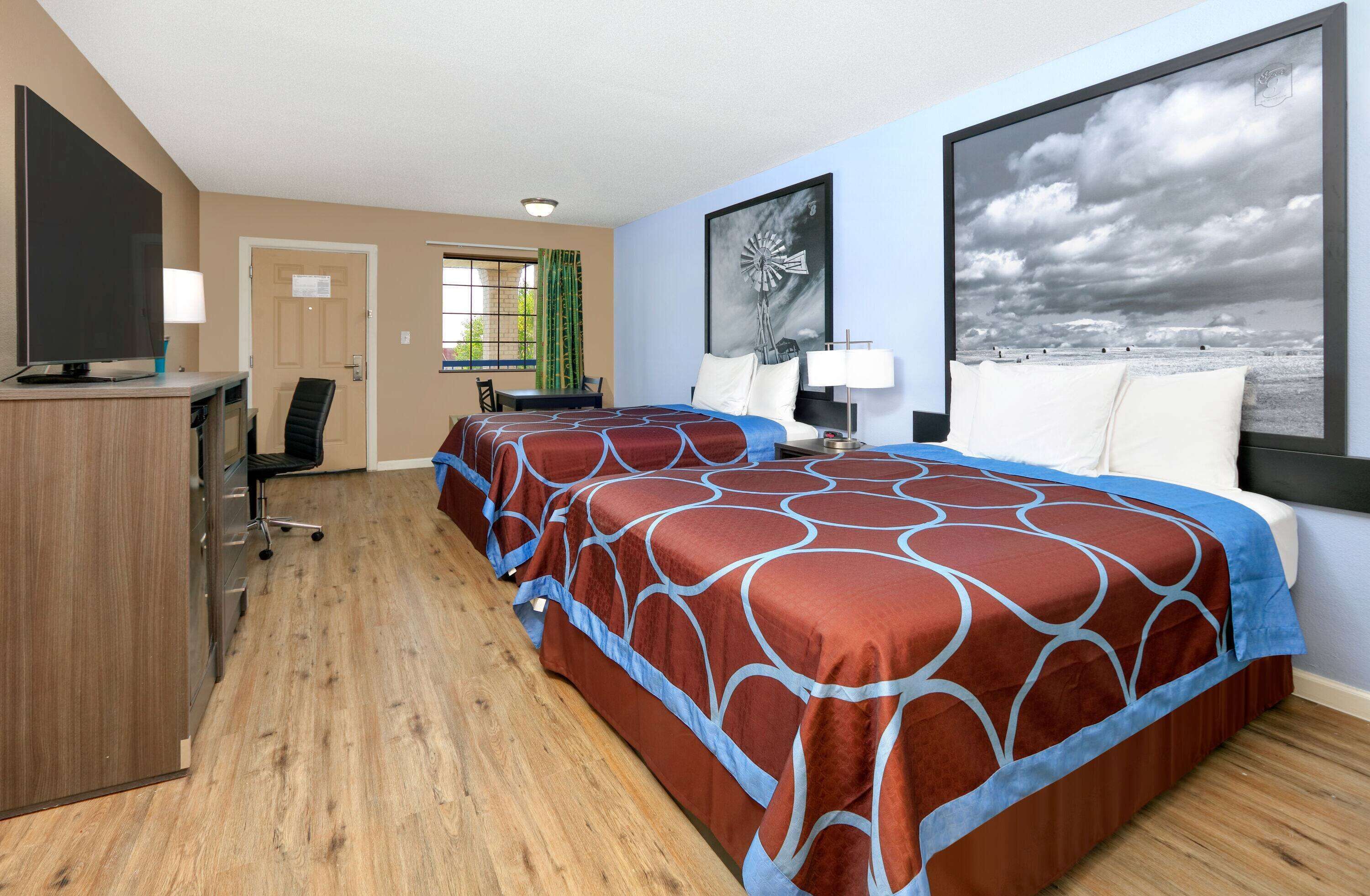 Habitación de hotel con dos camas, un escritorio, un televisor y un ventanal con vistas al cielo.