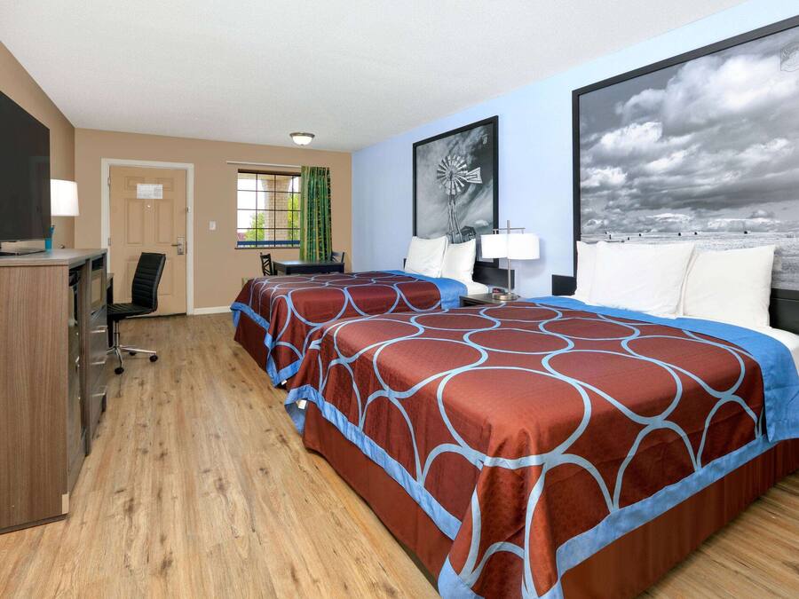 A hotel room with two beds, a desk, a TV, and a large window with a view of the sky.