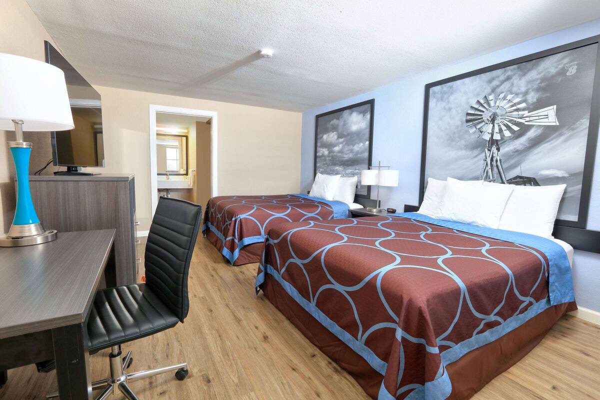 A hotel room with two beds, a desk, and a chair. There is a large window with a view of a windmill.