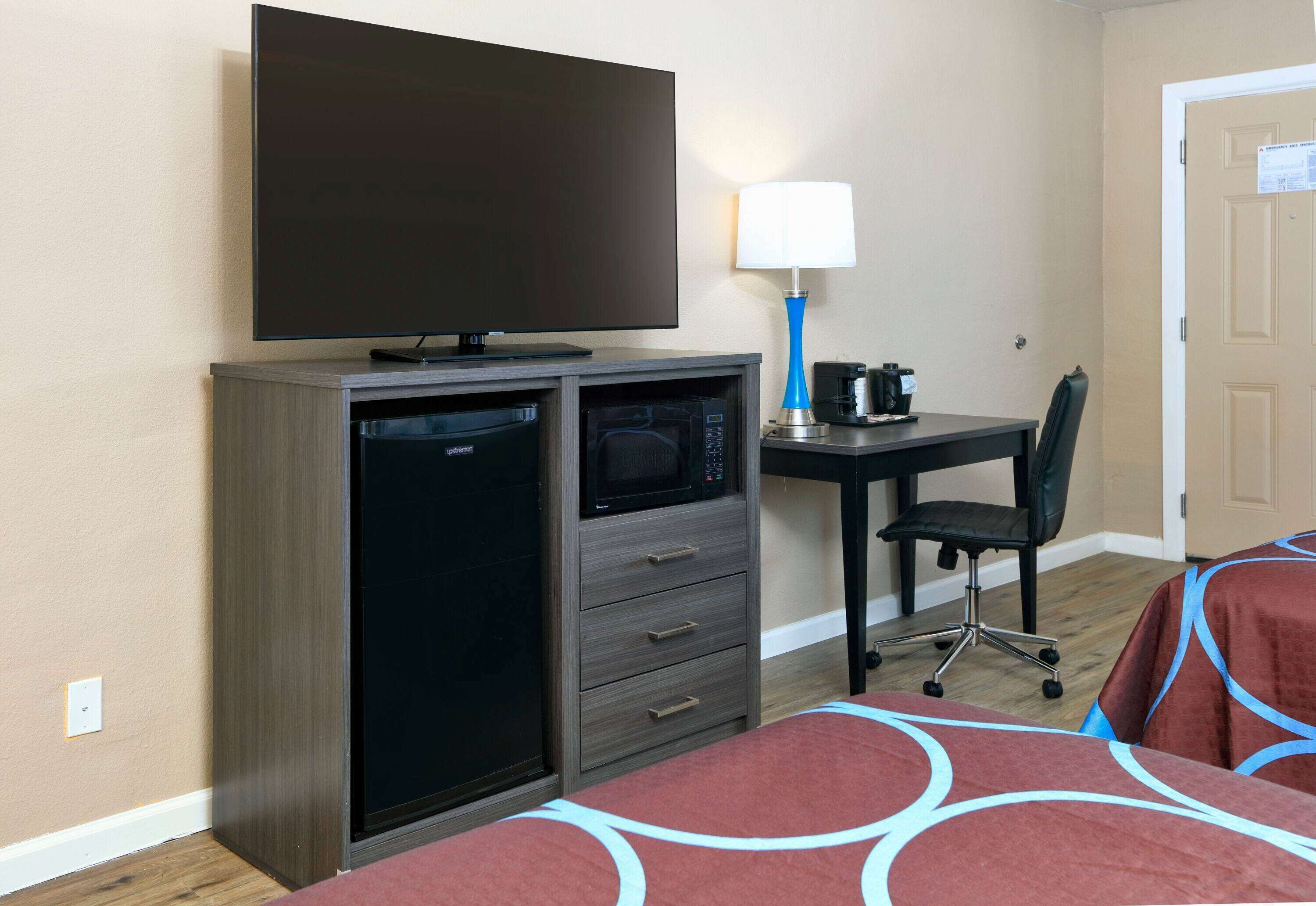 Habitación de hotel con televisor de pantalla plana, escritorio con lámpara, microondas, silla y cama con cubrecolchón estampado.