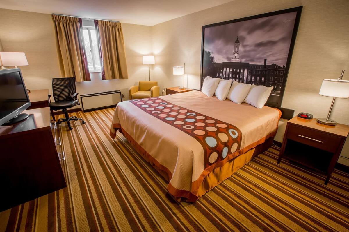 A hotel room with a bed, a desk with a computer, a chair, a lamp, and a painting on the wall.
