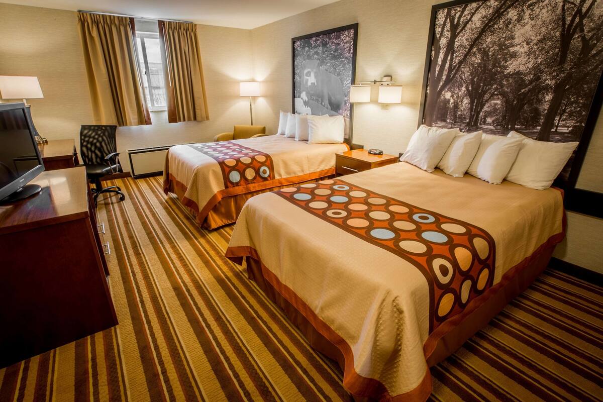 A hotel room with two beds, a desk, a TV, and a large window with curtains.