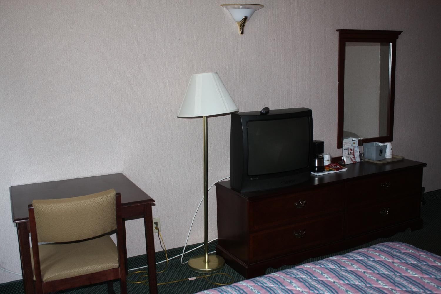 A hotel room with a bed, a desk, a chair, a television, a dresser, a lamp, and a mirror.