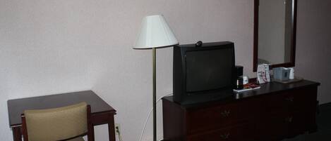 Una habitación de hotel con cama, escritorio, silla, televisor, cómoda, lámpara y espejo.