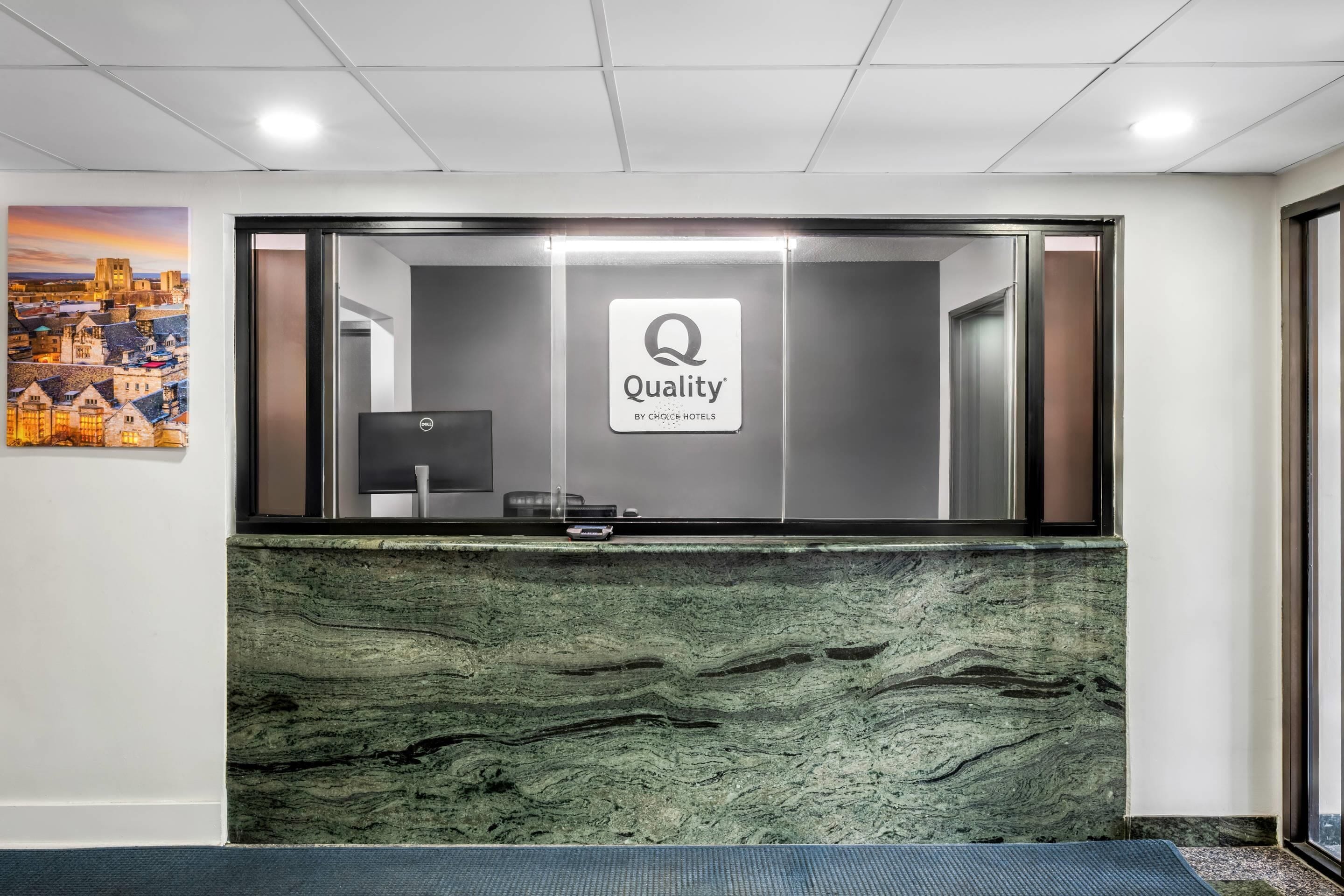 Una recepción con un cartel de 'Quality by Choice Hotels', una computadora y una pintura de un paisaje urbano en la pared.