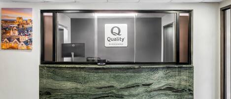 Una recepción con un cartel de 'Quality by Choice Hotels', una computadora y una pintura de un paisaje urbano en la pared.