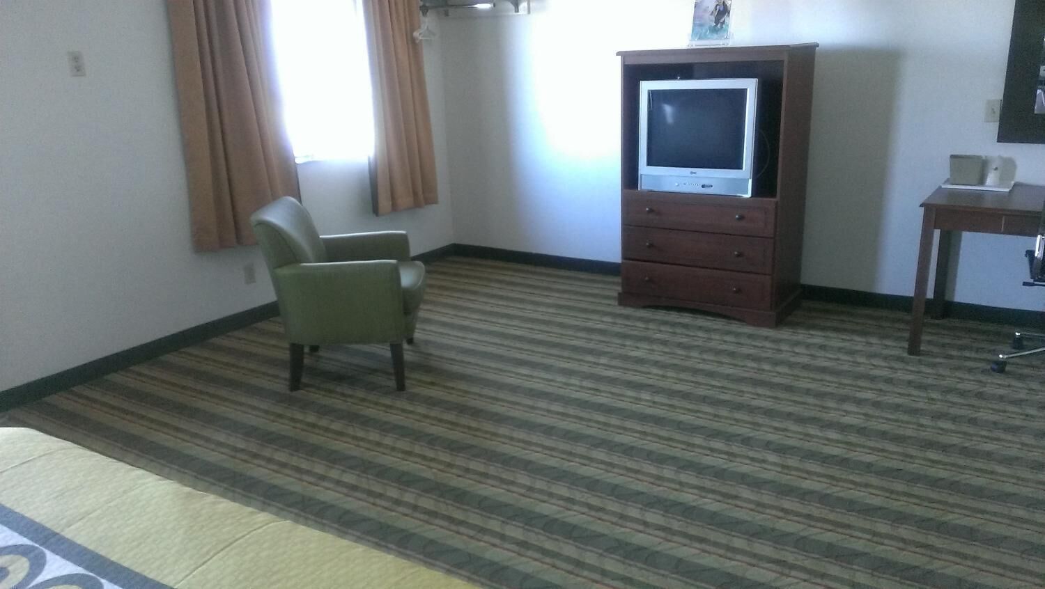 Habitación de hotel con alfombra a rayas, un sillón verde, un mueble de madera con televisor y una pequeña mesa con lámpara.