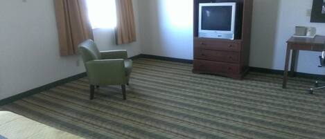 Habitación de hotel con alfombra a rayas, un sillón verde, un mueble de madera con televisor y una pequeña mesa con lámpara.