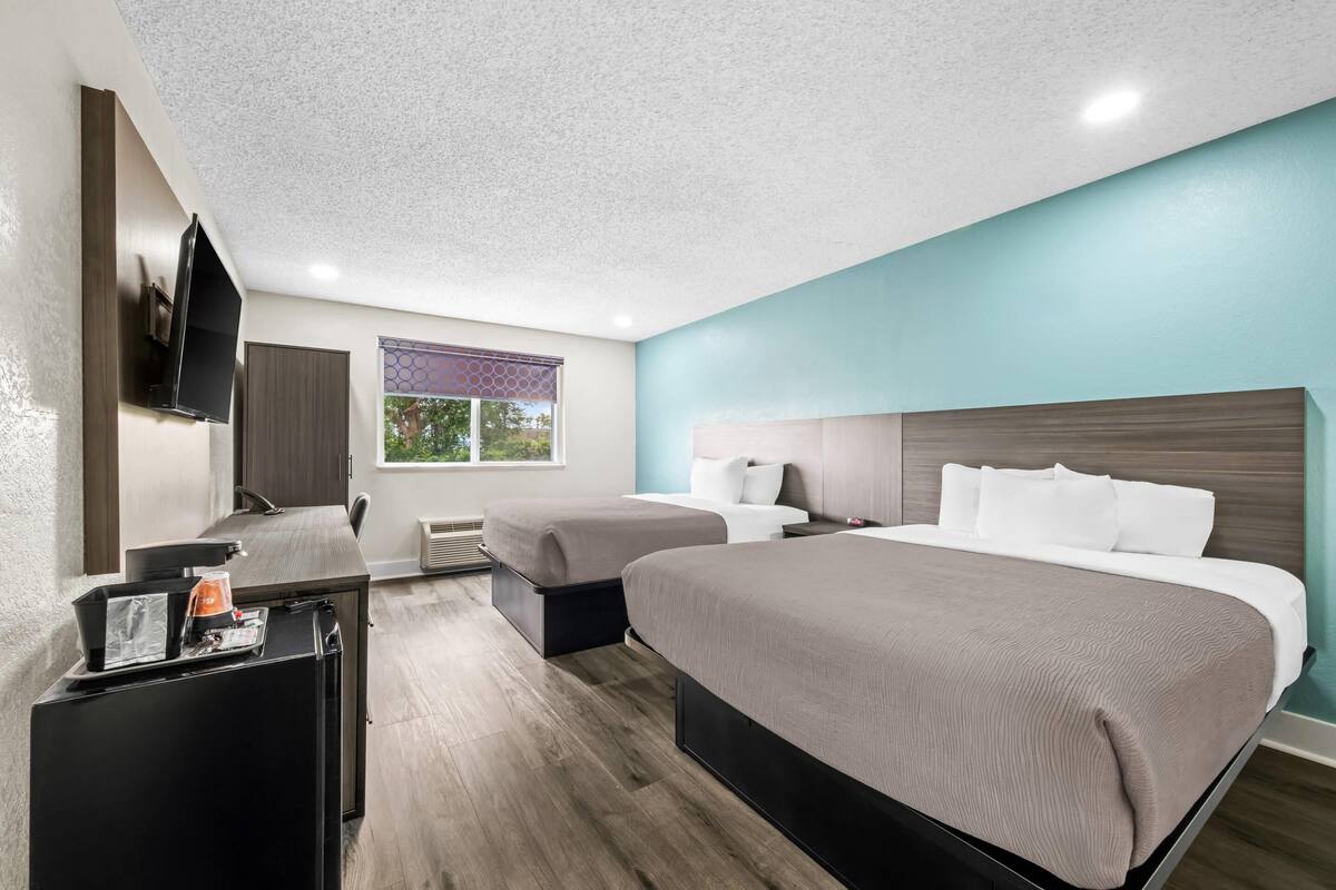 A hotel room with two beds, a desk with a coffee maker, a TV mounted on the wall, and a window with a view of trees.