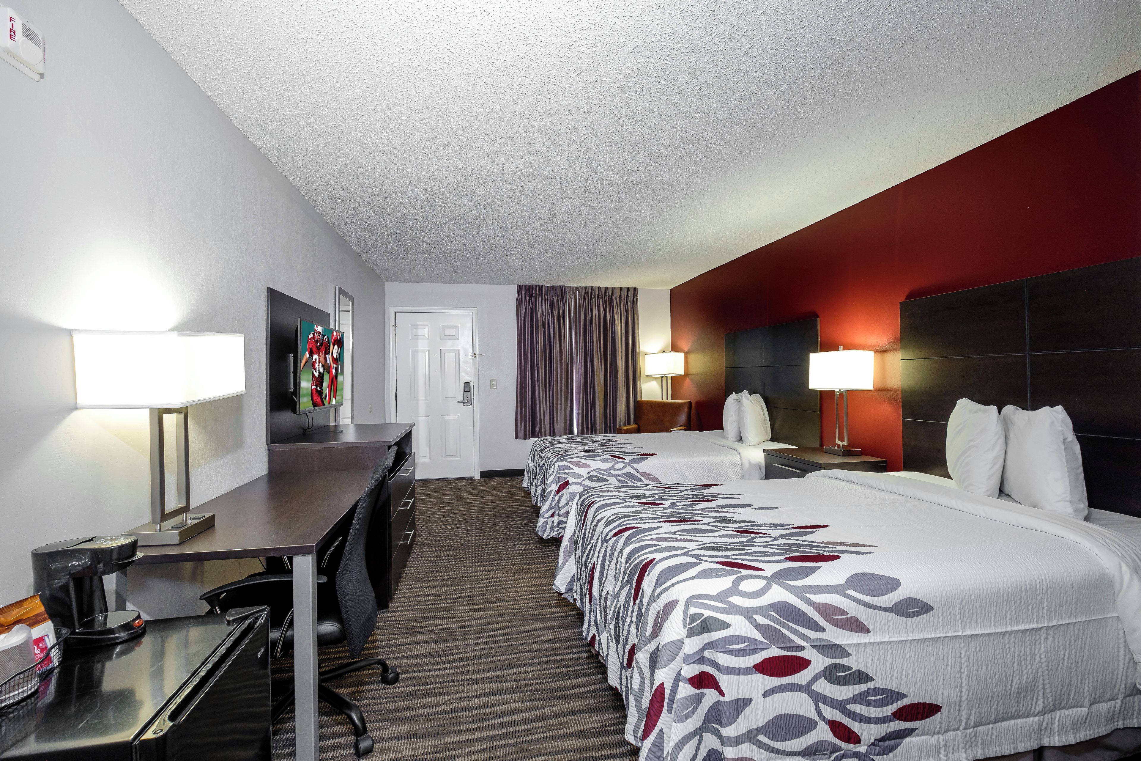 A hotel room with a long bed, a desk with a computer, a chair, and a TV on the wall.
