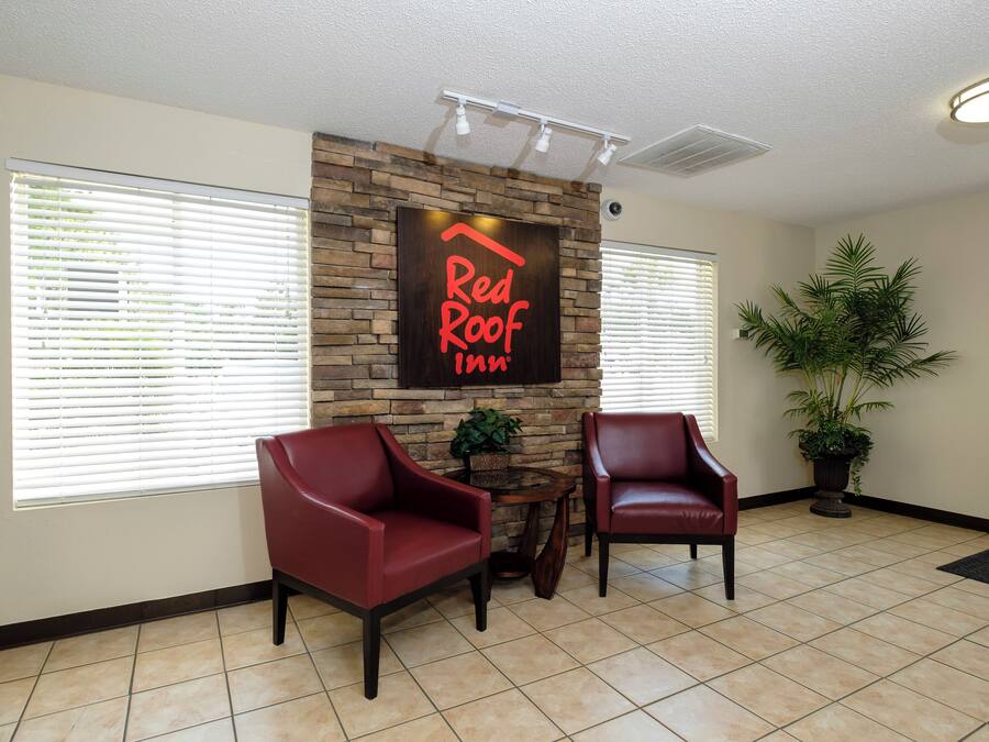 Un espace d’accueil avec une enseigne indiquant « Red Roof Inn » au-dessus d’un mur en pierre, deux fauteuils en cuir, une plante en pot et un sol carrelé.