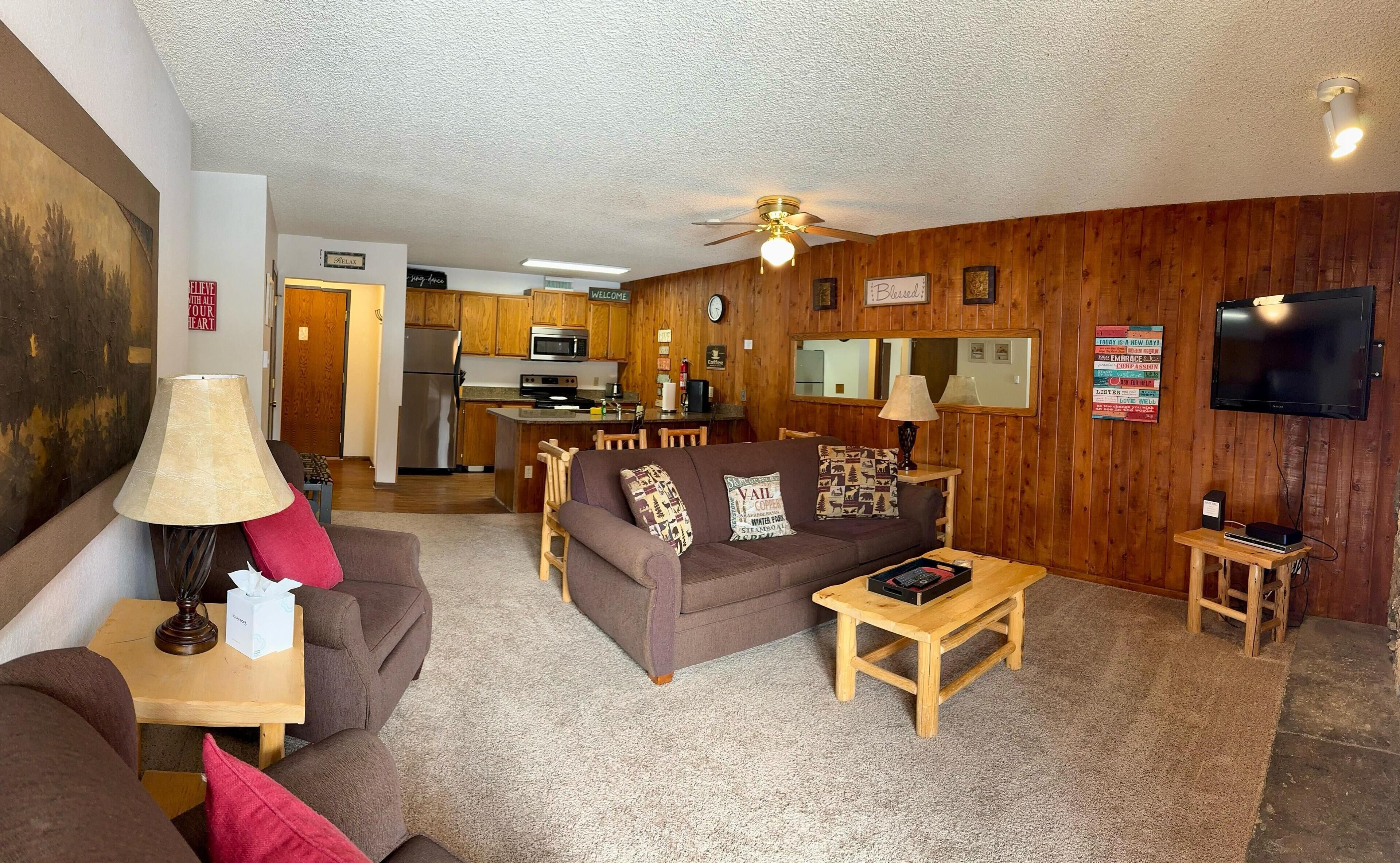 A cozy living room with a brown sofa, a coffee table, and a TV. There's a kitchen in the background with wooden cabinets and a refrigerator.