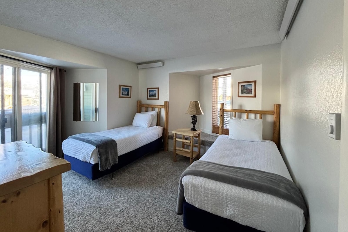 A hotel room with two single beds, a wooden desk, a lamp, and a window with curtains.
